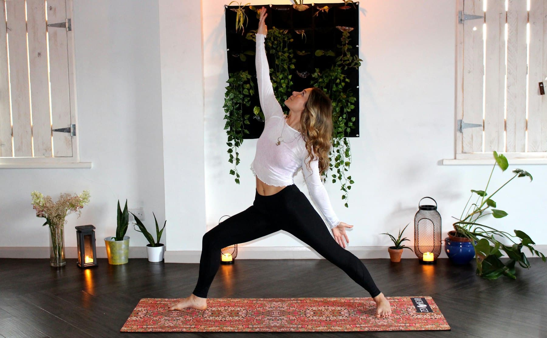 Woman in warrior pose on a patterned rug in a plant-filled studio